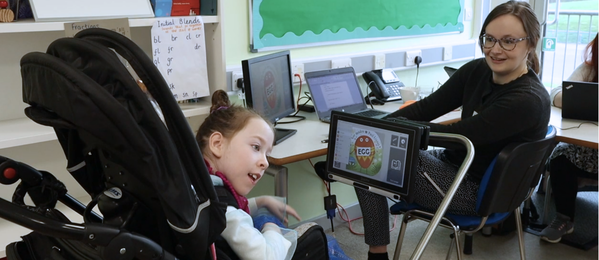 A student using Grid3 to read a book, she is smiling. There is a TA with her